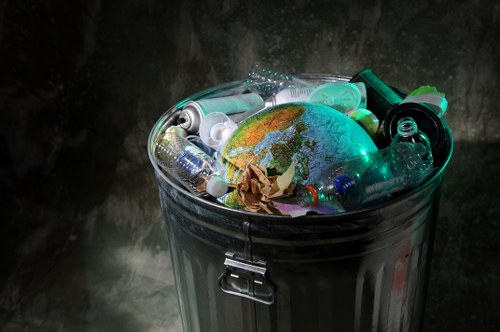 A metallic rubbish bin with a lid, positioned on a dark, reflective surface against a textured grey background, is filled with assorted discarded items including clear plastic bottles, some with labels and caps, crumpled brown paper, and a small globe depicting continents with blue oceans and green landmasses, which is partially visible amid the waste. The bottles vary in shape and size, with some caps attached and others loose, while the paper appears crushed and scattered among them. The globe sits prominently in the centre, contrasting with the surrounding plastic waste, emphasizing environmental themes related to rubbish and recycling. The scene is illuminated with soft, diffused lighting that highlights the reflective metallic surface of the bin and the varied textures of the waste items, creating a stark visual connection to rubbish removal and waste management services offered by Neasden House Clearance in London, with subtle references to local environmental challenges within the UK.
