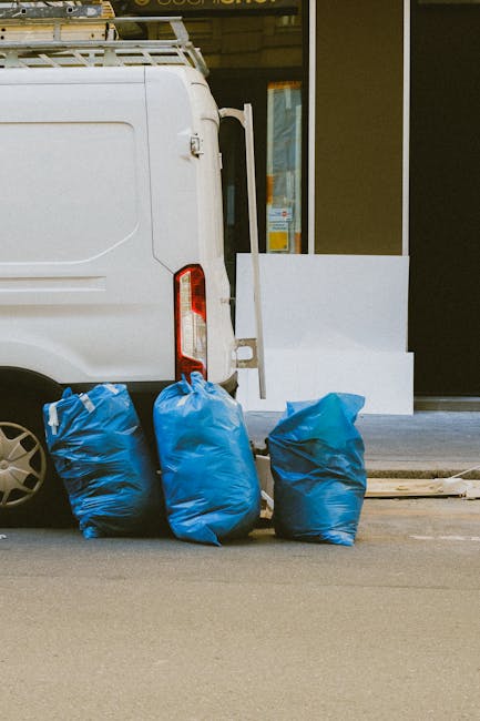 The image shows the rear section of a white commercial van parked on a street in front of a building with large glass windows and a dark exterior facade. Three heavy-duty blue rubbish bags, made of th