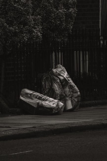 Two large black trash bags, made of shiny plastic material, are placed on the edge of a pavement in front of a black wooden fence with vertical slats and a lattice design. The bags are filled and tied